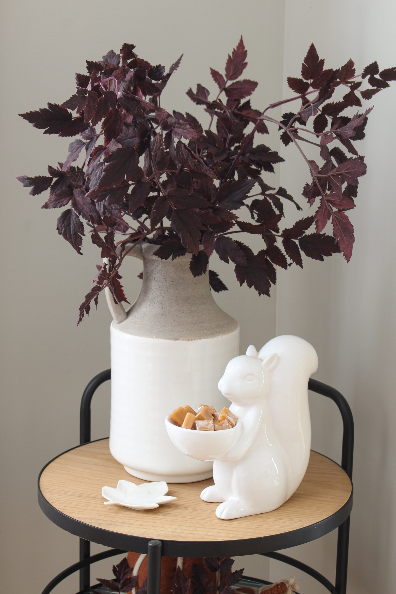 White ceramic squirrel holding candies on a tray with a vase of plum cimicifuga stems.