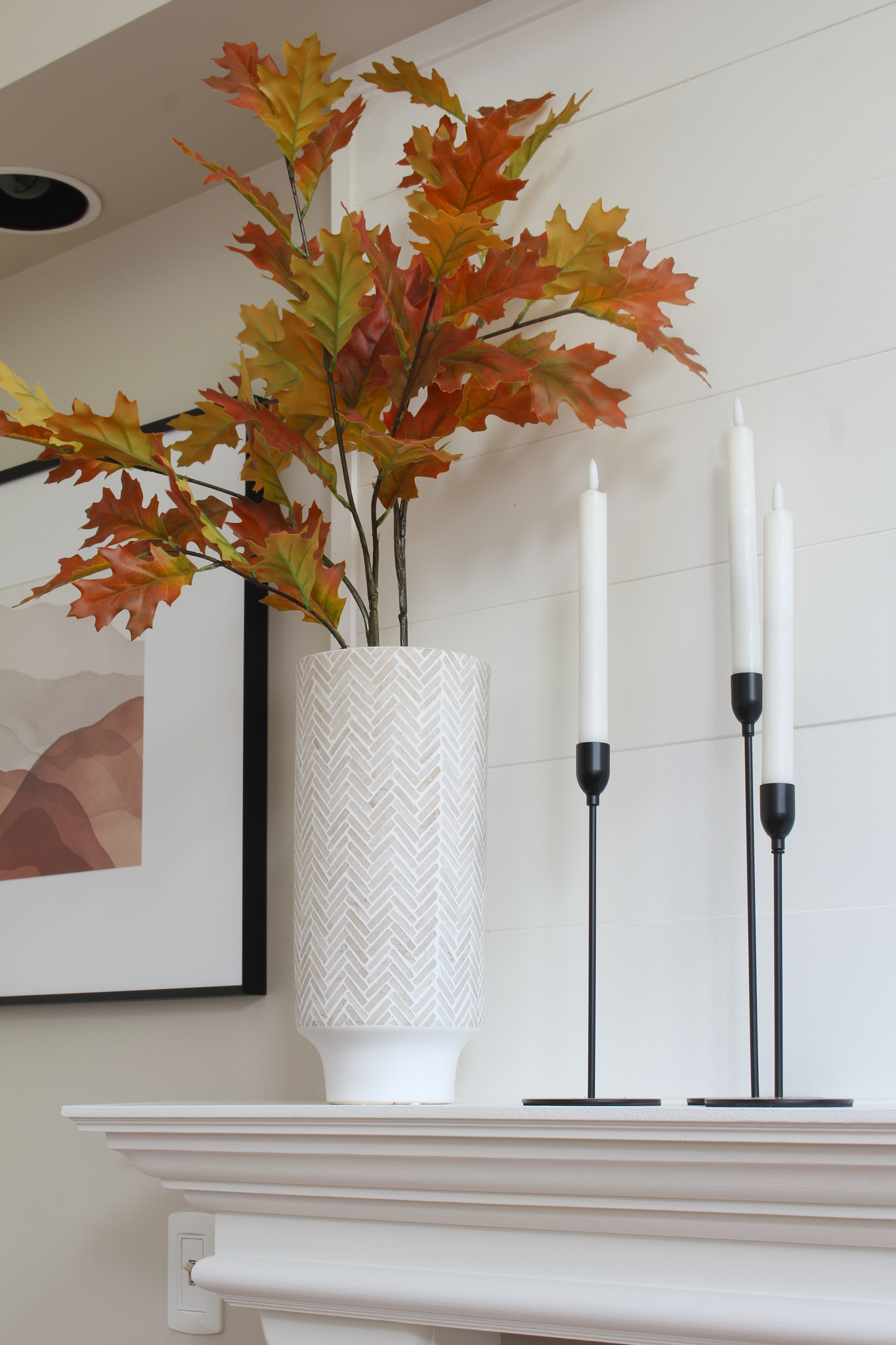 Vase with colorful oak leave with black candlesticks on a white mantel.