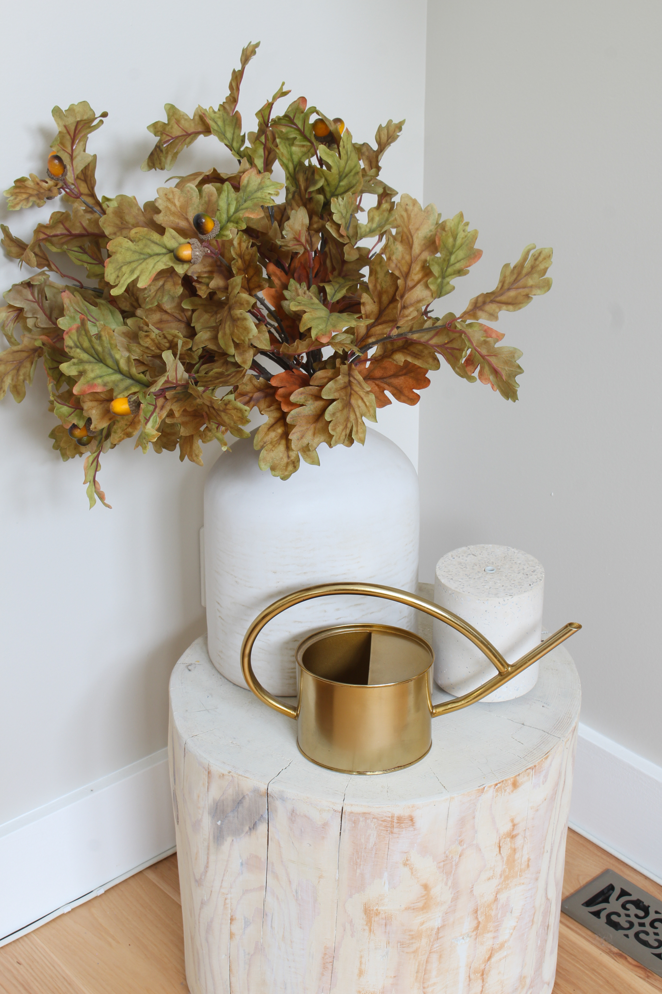 Vase with oak leaves, diffuser, and gold watering can on a wood stump side table.