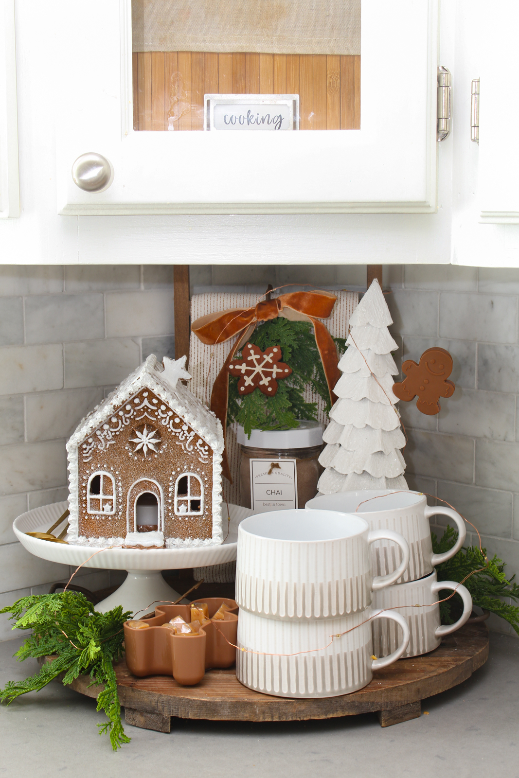 Round wood kitchen tray decorated with a lighted gingerbread house, mugs, and a tree for a cute Christmas display.