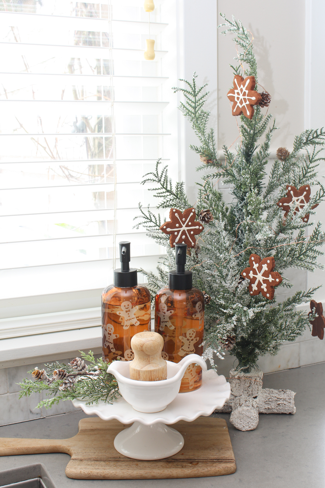 Christmas sink display with gingerbread men soap dispensers on a white cake stand with a small tree in the background.