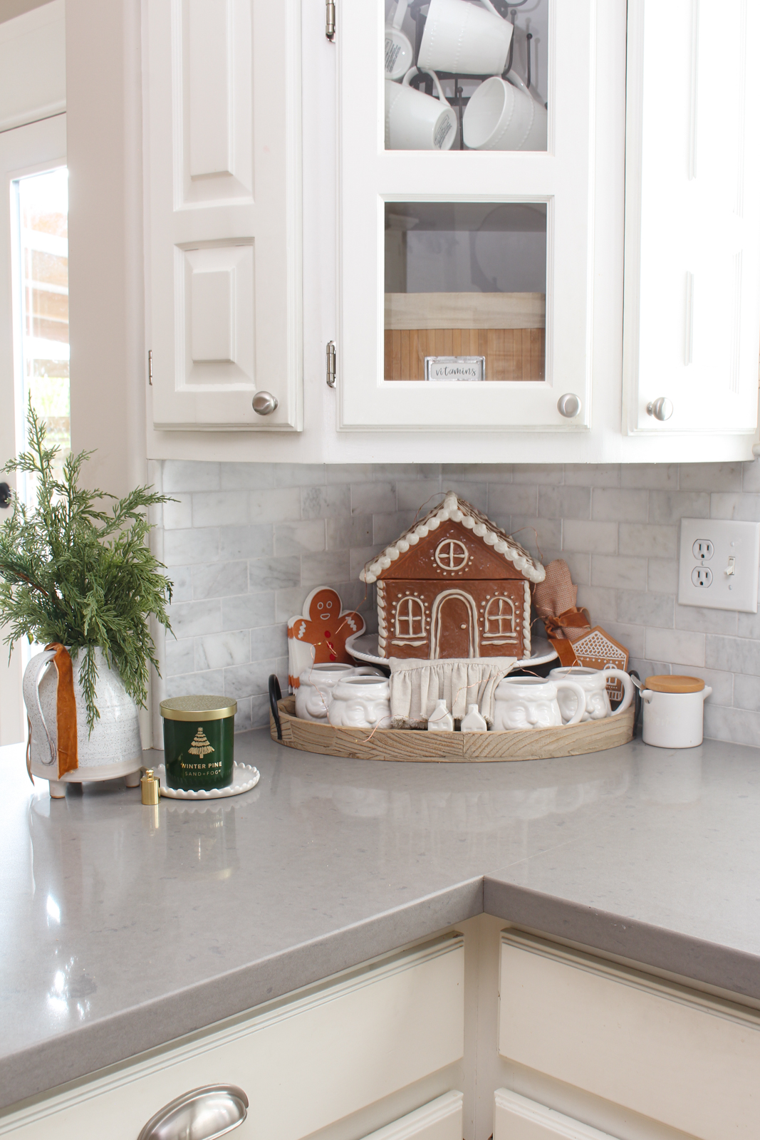 Gingerbread house cookie jar on a tray with white Santa mugs in a white kitchen. 