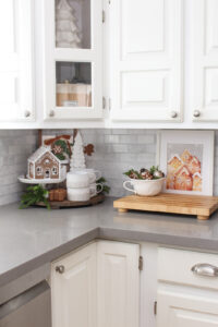 White kitchen with pretty gingerbread decor. A round tray is decorated in the corner with a gingerbread house and white tree. A white bowl with faux gingerbread decorations and a gingerbread village print are on the counter.