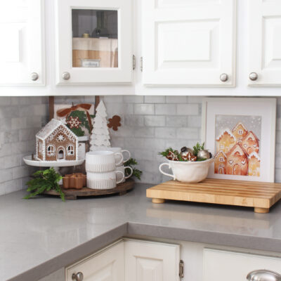 White kitchen with pretty gingerbread decor. A round tray is decorated in the corner with a gingerbread house and white tree. A white bowl with faux gingerbread decorations and a gingerbread village print are on the counter.