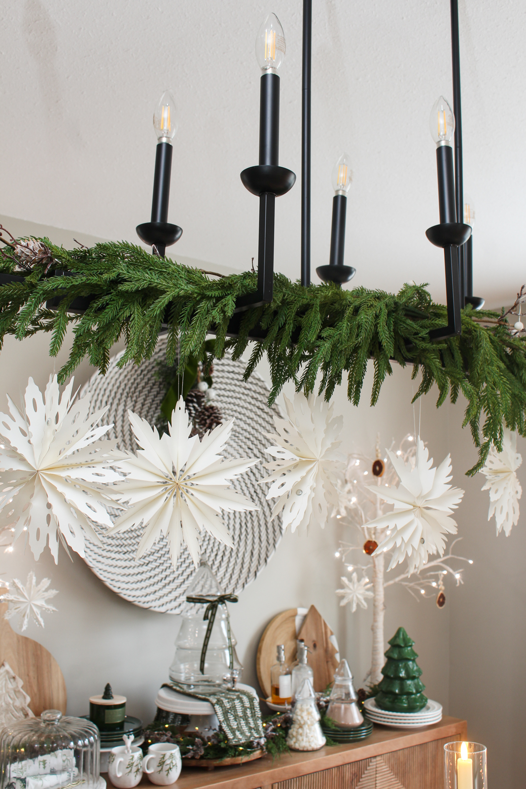 Black dining room light fixture decorated for Christmas with faux greenery, a twig garland, and paper snowflakes.