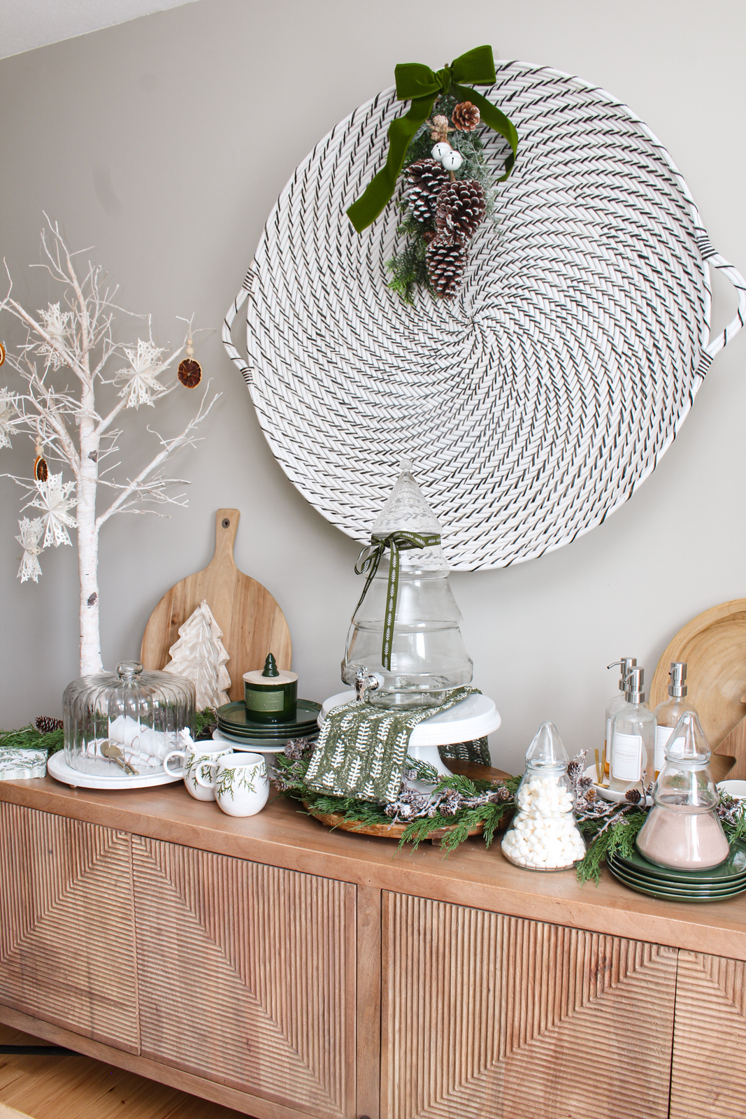 Wood sideboard decorated for Christmas with Christmas tree glass beverage dispenser and hot chocolate bar.