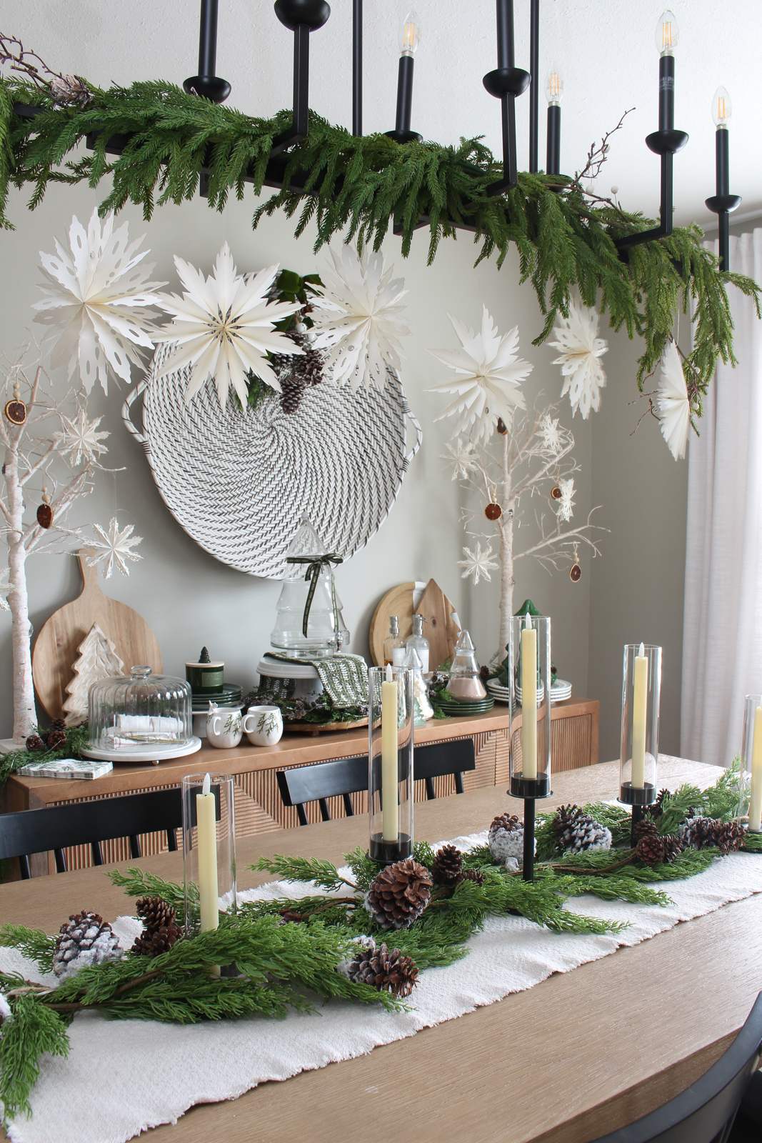 Christmas dining room with centerpiece using black candle sticks, faux greenery, snowy pinecones, and lights.