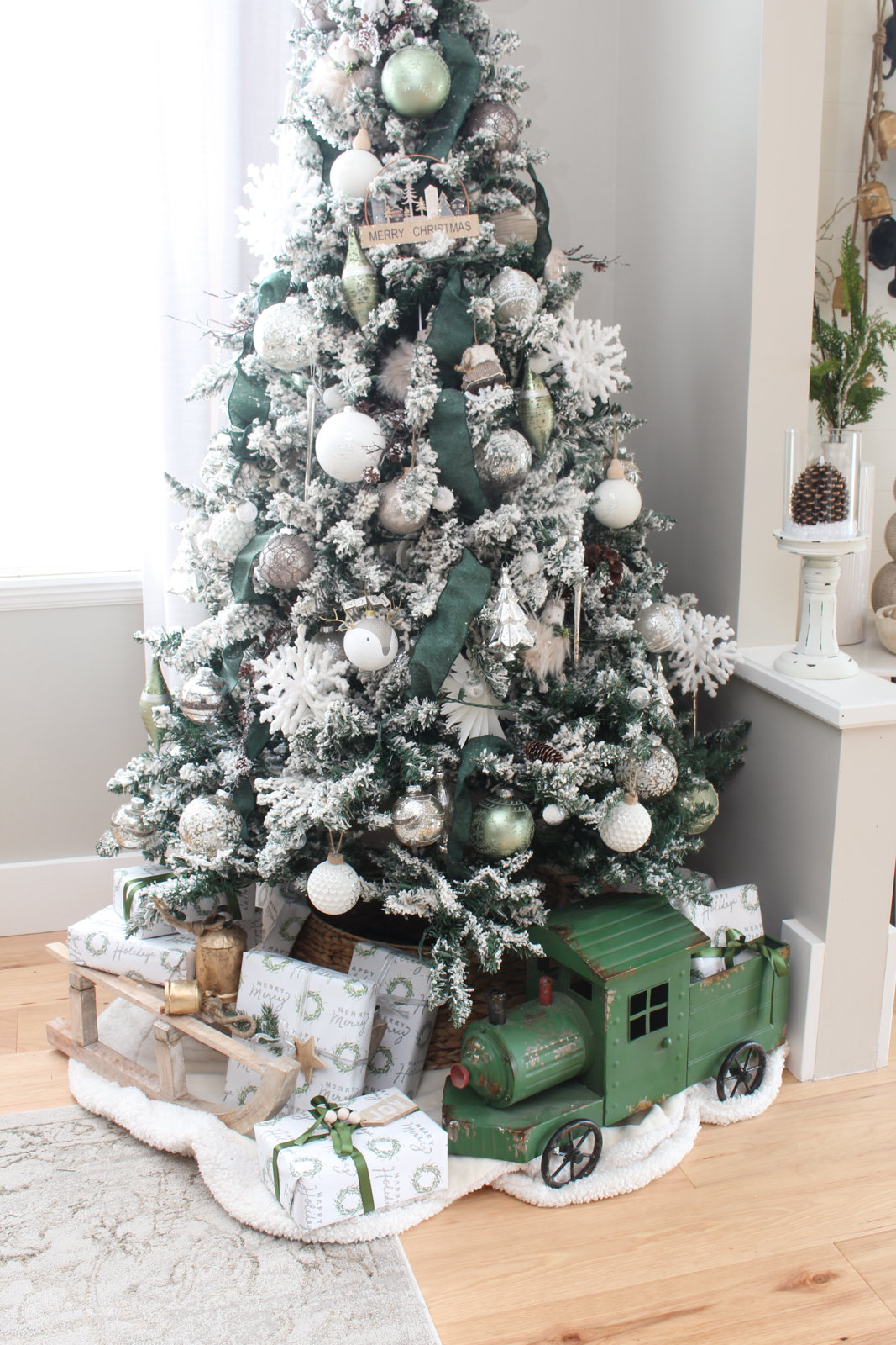 Christmas dining room Christmas tree decorated with a winter wonderland theme. Flocked tree is filled with green riboon, silver and white decorations, and woodsy touches just as branches and pinecones.