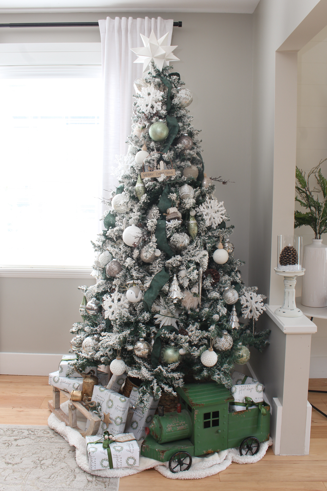 Christmas dining room Christmas tree decorated with a winter wonderland theme. Flocked tree is filled with green riboon, silver and white decorations, and woodsy touches just as branches and pinecones.